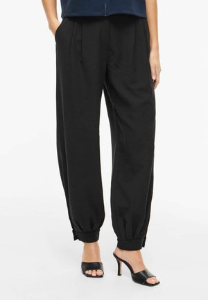 Zwarte loszittende broek met omslag bij de enkels, gedragen met zwarte open-toe hoge hak mules, handen in de zakken, tegen een witte achtergrond.