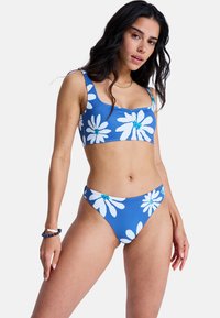 Σετ μπικίνι από μπλε ύφασμα με μεγάλα λευκά σχέδια μαργαρίτας. Το πάνω μέρος έχει τετράγωνη λαιμόκοψη· το κάτω μέρος έχει χαμηλή μέση.