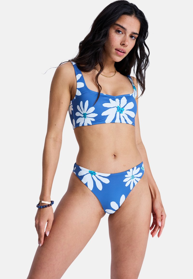 Σετ μπικίνι από μπλε ύφασμα με μεγάλα λευκά σχέδια μαργαρίτας. Το πάνω μέρος έχει τετράγωνη λαιμόκοψη· το κάτω μέρος έχει χαμηλή μέση.