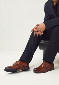 Chaussures habillées en cuir marron avec détails brogues, design à lacets, bout arrondi, et texture lisse sur un fond noir et blanc.