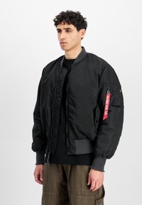 Čierna bomber bunda s rebrovými manžetami, predným zipsom a vreckom na ľavom rukáve s červenou nášivkou. Hladká, ľahká tkanina.
