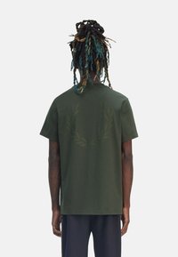 Camiseta de algodón verde oliva de corte regular, con un discreto diseño de una corona de olivo en la parte posterior y mangas cortas. Sin marcas visibles.