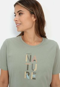 Lichtgroene katoenen t-shirt met een ronde halslijn en korte mouwen, voorzien van een geprinte slogan "EMPOWERED BY NATURE" in verschillende kleuren.