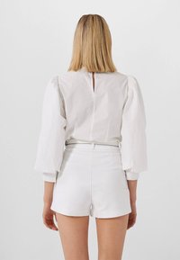 Bílý top s nabíranými rukávy a malým klíčkovým výřezem na zádech. V kombinaci s bílými šortkami s vysokým pasem. Hladká textura a minimalistický design.