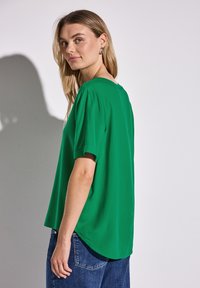Blusa verde realizzata in tessuto leggero con maniche corte, scollatura rotonda e orlo posteriore leggermente allungato, abbinata a jeans blu.