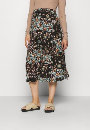 Woman wearing a midi-length black floral skirt with patchwork design, beige long-sleeve top, and beige slip-on sandals with black soles.