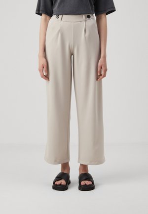Beige wijde broek met een gladde textuur, voorplooien en twee zwarte knopen in de taille, gecombineerd met zwarte schuifsandalen.