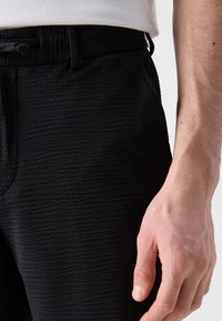 Pantaloni negri texturati cu un model de grilă subtil, având buzunare laterale și un cordon la talie, ținuți în mâna unei persoane.