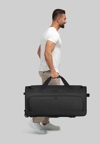 Hombre con camiseta blanca y pantalones beige llevando una gran bolsa de deporte negra con ruedas por las asas, contra un fondo gris.