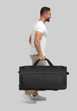 Uomo in t-shirt bianca e pantaloni beige che porta una grande borsa da viaggio nera con ruote per le maniglie su sfondo grigio.