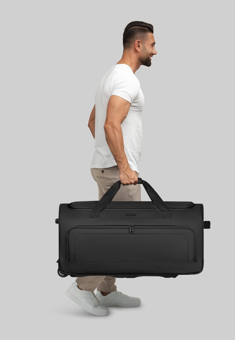 Hombre con camiseta blanca y pantalones beige llevando una gran bolsa de deporte negra con ruedas por las asas, contra un fondo gris.