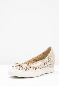 Chaussure beige à talon compensé perforé avec semelle blanche et nœud décoratif à l’avant, vue de côté sur fond blanc.