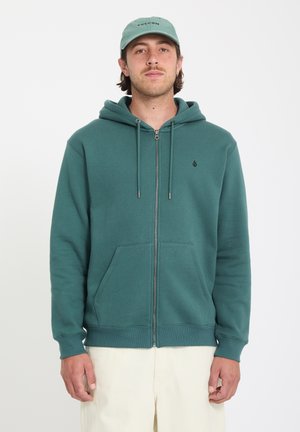 Młody mężczyzna w turkusowej bluzie na zamek z przednimi kieszeniami i jasnooliwkową czapką, stojący na białym tle.