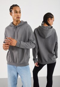 Felpa con cappuccio grigia, dotata di una comoda tasca frontale, polsini e orlo a coste. Il modello mostra una vestibilità rilassata; il tessuto appare morbido e liscio.