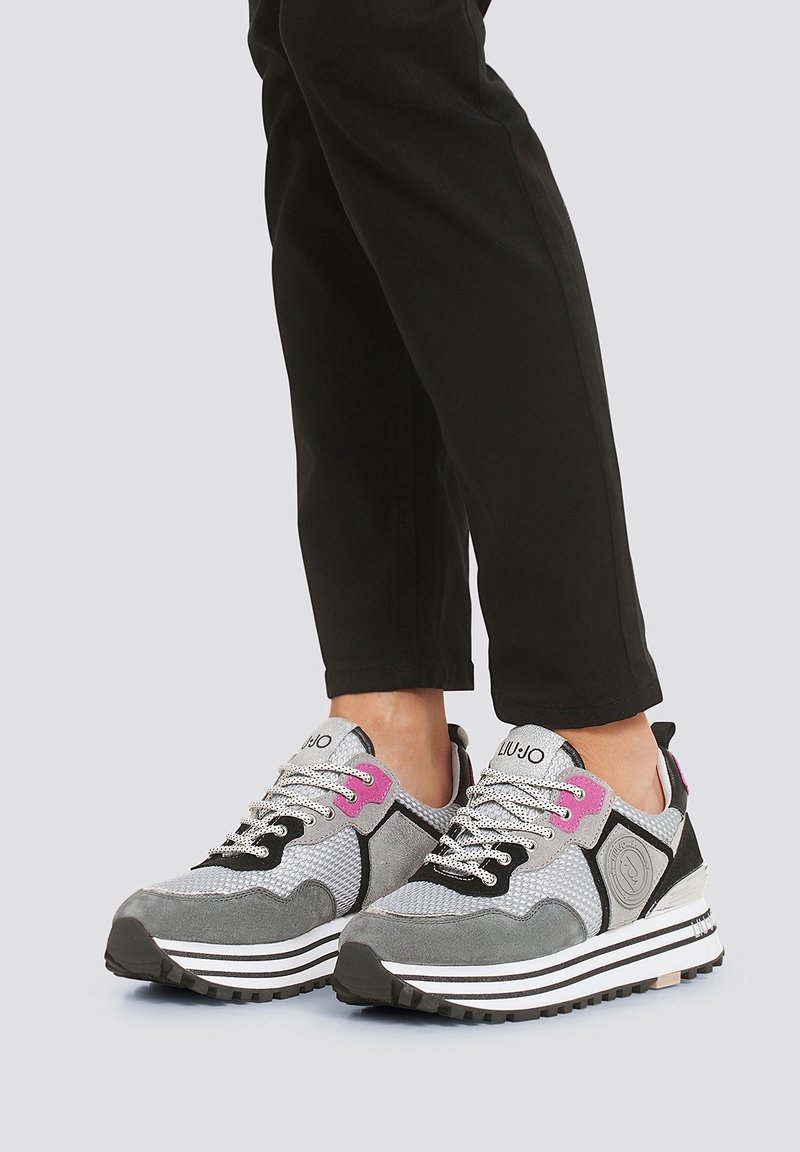 Personne portant des baskets plateformes gris, noir, blanc et rose, avec des lacets à motifs et un pantalon noir, sur un fond gris.