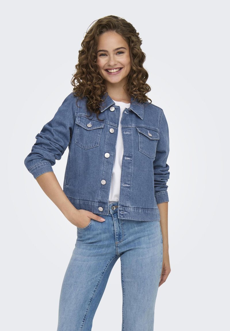 Veste en denim bleu avec deux poches poitrine, boutons argentés et design ajusté. Portée sur une chemise blanche, assortie à un pantalon en jean bleu.