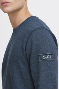 Sudadera azul oscuro con cuello redondo, textura suave y una pequeña etiqueta bordada "Solid" en la manga izquierda. Diseño minimalista.