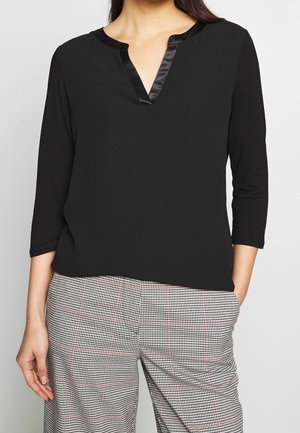 Femme portant un haut noir à manches longues avec un décolleté en V satiné et un pantalon à carreaux noir et blanc avec des lignes rouges.