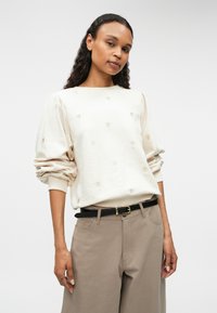 Bežasti pulover s okruglim izrezom i napuhanim rukavima. Ima zlatnu vezenu aplikaciju u obliku srca, u kombinaciji s labavim smeškim hlačama i crnim remenom.