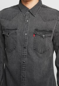 Camicia in denim grigio con colletto a punta, due tasche sul petto e cuciture a contrasto. Presenta una piccola etichetta rossa sul taschino.
