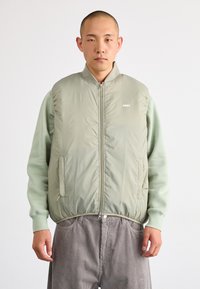 Helle grüne Steppweste mit Reißverschluss vorne, Seitentaschen und elastischem Saum. Getragen über einem hellgrünen Sweatshirt und kombiniert mit hellgrauen Cordhosen.