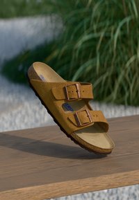 Bruna mockaskinnsandaler med två justerbara bronzefärgade spännen, formad fotbädd och texturerad sula, placerade på en träytan med en grönskande bakgrund.