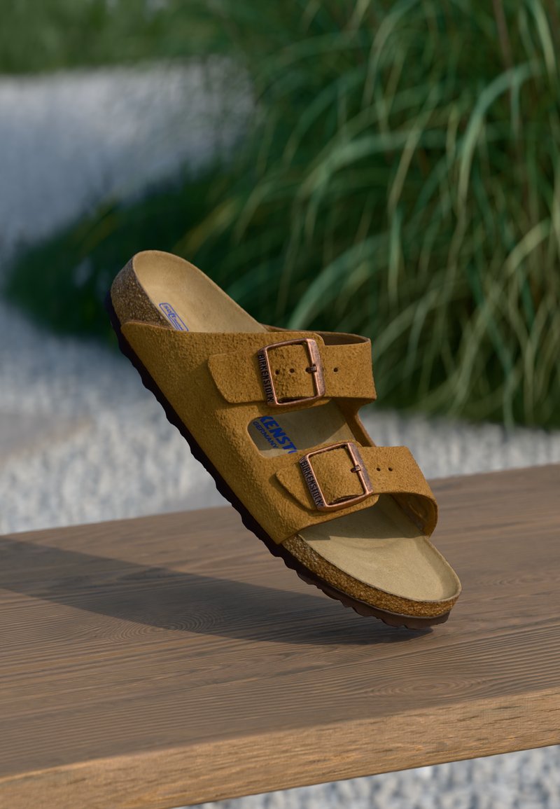 Bruna mockaskinnsandaler med två justerbara bronzefärgade spännen, formad fotbädd och texturerad sula, placerade på en träytan med en grönskande bakgrund.
