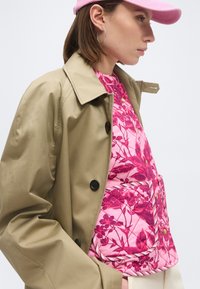 Γυναίκα με μπεζ καμπαρντίνα πάνω από ροζ φλοράλ καπιτονέ γιλέκο και ροζ καπέλο μπέιζμπολ, απεικονίζεται σε προφίλ μπροστά σε λευκό φόντο.