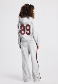 Grauer Hoodie und Jogginghosen-Set, mit großem maroonfarbenen "89" Druck auf dem Rücken und maroonfarbenen Seitestreifen an der Hose. Weicher Stoff, lässige Passform.