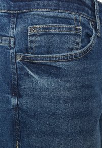 Jeans de ganga azul escuro com detalhe no bolso traseiro, costura reforçada e ligeira descoloração no lado superior esquerdo.