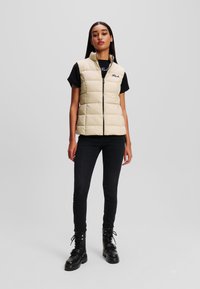 Beige Puffer-Weste mit einem Reißverschluss vorne, genähten Quadraten und einem Stehkragen, kombiniert mit einem schwarzen T-Shirt, engen Jeans und schwarzen Schnürstiefeln.