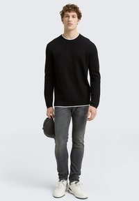 Schwarzer gerippter Pullover mit langen Ärmeln, Akzent in Weiß am Rundhalsausschnitt, kombiniert mit dunkelgrauen Jeans und weißen Turnschuhen, hält eine graue Kappe.