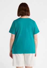 Camiseta de manga corta en color verde azulado con cuello redondo, textura suave y corte relajado. La parte posterior presenta una costura sencilla y sin estampados.
