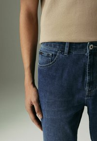 Jeans din denim albastru au un design clasic cu cinci buzunare, având detalii de cusătură subtile. Materialul are o textură netedă și zone ușor decolorate.