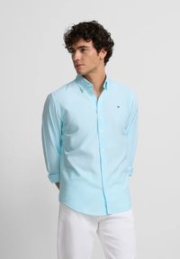 Hombre de pie con las manos detrás de la espalda, vistiendo una camisa de botones azul clara con las mangas arremangadas y pantalones blancos, frente a un fondo liso.