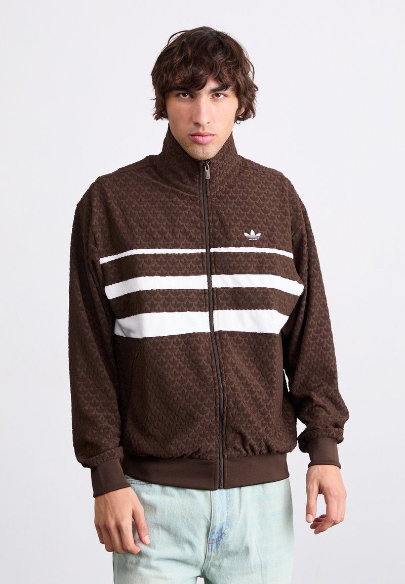 Καφέ ζακέτα με φερμουάρ με υφή υφάσματος, διαθέτει λευκές ρίγες σταυρωτά στο στήθος και ψηλό γιακά. Διακοσμημένη με ένα μικρό λογότυπο στο αριστερό στήθος.