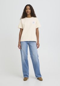 Béžna bombažna majica s kratkimi rokavi in modrim cvetličnim motivom na levi strani prsnega koša. Kombinirana s svetlo modrimi hlačami z ravnimi hlačnicami.