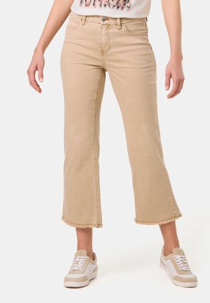 Beige cropped jeans with a frayed hem, featuring a classic five-pocket design and a zip and button closure. Paired with white sneakers.