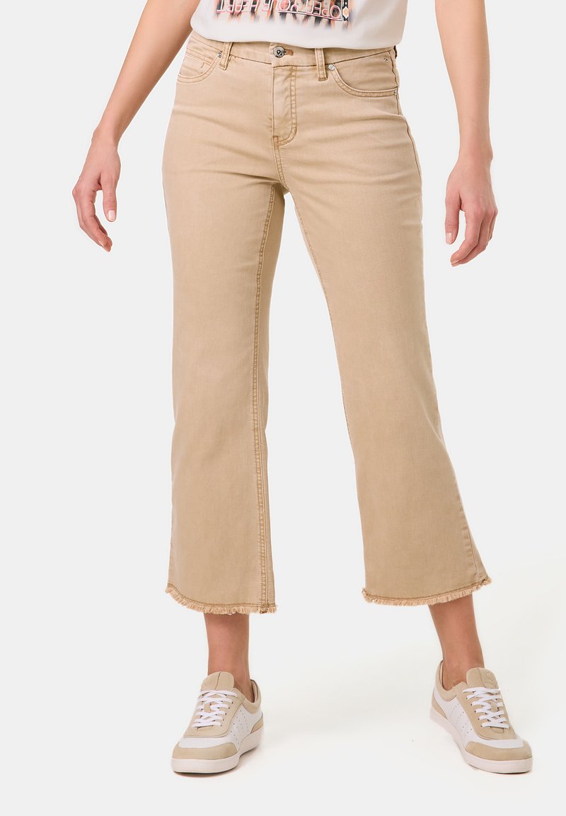 Beige cropped Jeans mit fransigem Saum, klassischem Five-Pocket-Design und einem Reiß- und Knopfverschluss. Kombiniert mit weißen Sneakers.