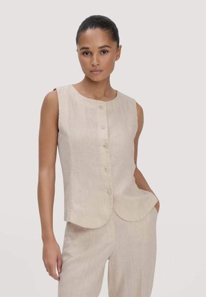 Frau trägt ein ärmelloses, beigefarbenes, geknöpftes Oberteil und passende Hose, mit einer Hand in der Tasche, steht vor einem schlichten Hintergrund.