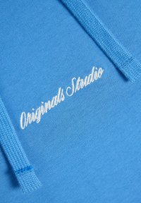 Sinine kapuutsiga, millel on nähtav tekstuuriline kangas, valge telgedega "Originals Studio" logo ja sobivad sinised nöörid.