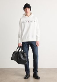 Felpa bianca con logo GANT 1949, jeans blu, stivali neri e una grande borsa da viaggio nera. Il modello indossa un berretto nero e sta in piedi.