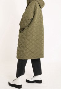 Olijfgroene, gewatteerde pufferjas met een capuchon, zakken en een middellange pasvorm, gecombineerd met zwarte jeans en witte veterschoenen.