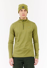 Man in olijfgroene half-zip pullover en bijpassende beanie met logo's, staand tegen een effen witte achtergrond.