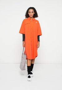 Abito camicia arancione oversize con bottoni frontali, maniche corte e tasca sul petto. Abbinato a una borsa grigia e calzini neri alla caviglia.