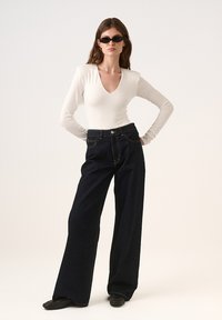 Biały body z długim rękawem i głębokim dekoltem w kształcie V, zestawione z szerokimi, ciemnoniebieskimi dżinsami z żółtym przeszyciem i zapięciem na guzik. Czarne okulary przeciwsłoneczne.