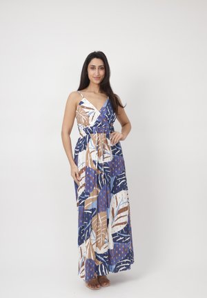 Donna in piedi con una mano sui fianchi, indossa un abito lungo senza maniche con motivi astratti di foglie in blu, marrone e bianco.