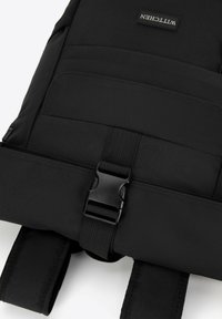 Mochila negra con un exterior de tela elegante, cierre de hebilla, correas de hombro acolchadas y detalles de costura visibles.