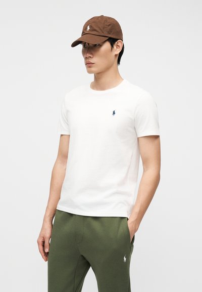 Άνδρας που φορά καφέ καπέλο, λευκό κοντομάνικο πουκάμισο και πράσινο παντελόνι με λογότυπα, στέκεται με το ένα χέρι στην τσέπη μπροστά σε ένα απλό φόντο.