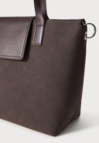 Brun toteveske med teksturert finish, med en glatt lærklaff, kontraststing, og en sølvringdetalj på siden.